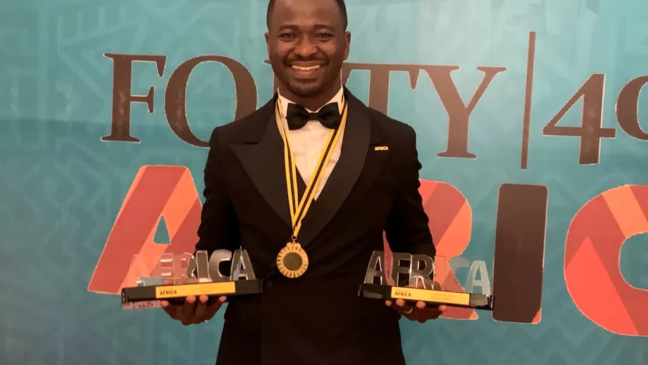 A person in a tuxedo holds two awards in front of a banner that reads FORTY | 40