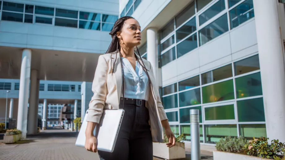 A woman walks away from building in suit with laptop